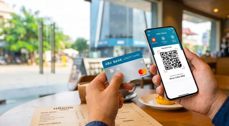 Please create an image with these image with prompt below, and a man are holding a Bank credit card in his hand: A first-person point-of-view shot of a modern, sleek smartphone screen displaying a bright, secure digital mobile banking app interface with a visible QR payment screen. The background is a beautifully blurred, warm-toned upscale cafe interior in Phnom Penh, Cambodia, with soft natural lighting streaming through large glass windows. Professional corporate aesthetic, clean layout, photorealistic, commercial style photography, high depth of field. --ar 16:9
